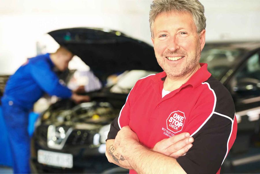 A smiling One Stop Check technician in the branded red polo, with a mechanic working under a bonnet behind
